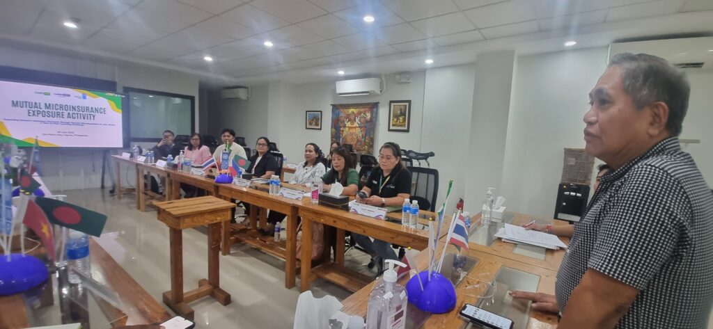A group of people sit at a conference table with country flags, listening to a presenter. A screen displays “Mutual Microinsurance Exposure Activity” during the UNDP IRFF Asia Pacific Peer Microinsurance Exchange in a bright meeting room.
