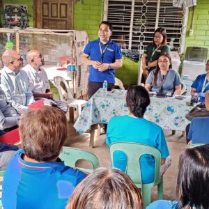 A group of people sits in a semi-circle on plastic chairs, attentively listening to a speaker at what appears to be an informal community meeting discussing the ICMIF Foundation and Microinsurance Network's global initiative to map community-based microinsurance.